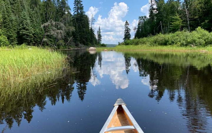 In the foreground, the tip of a canoe appears above very calm water, reflecting the grasses and trees on the shore, and the blue sky and clouds above. 